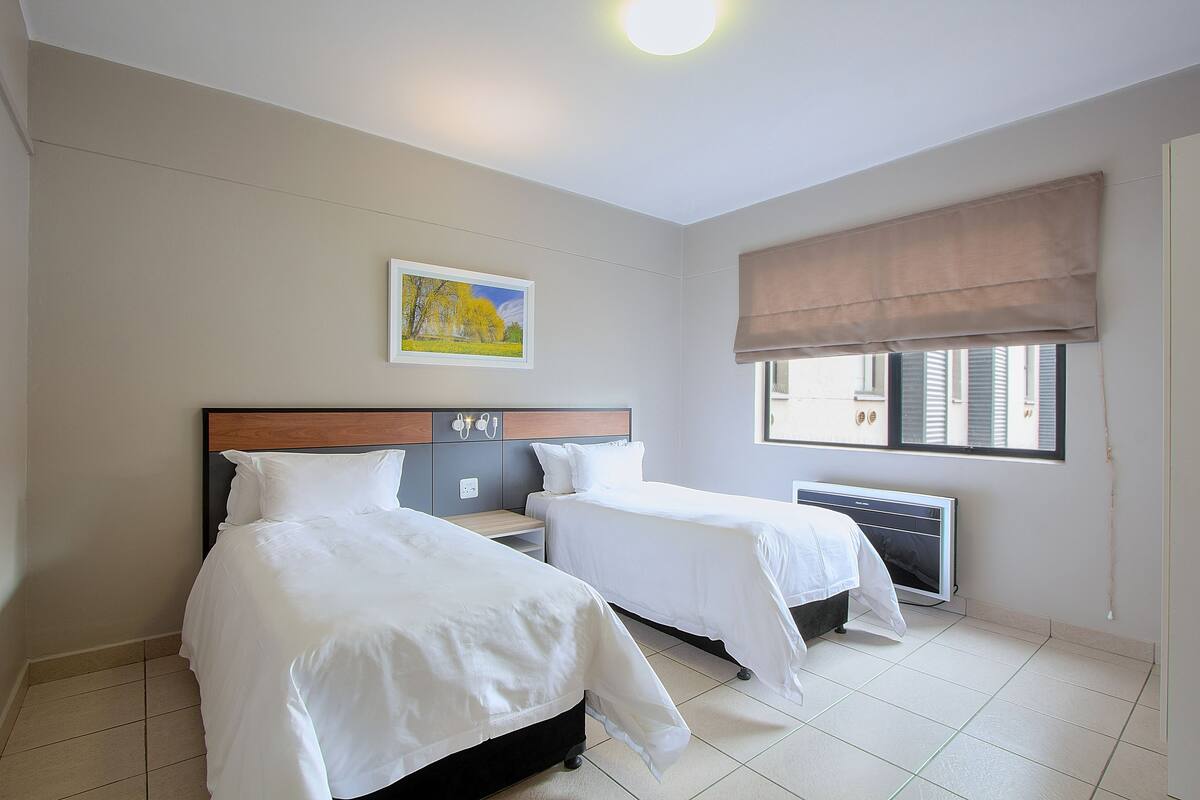 A hotel room with two beds, a nightstand, a radiator, and a window with a view of buildings.