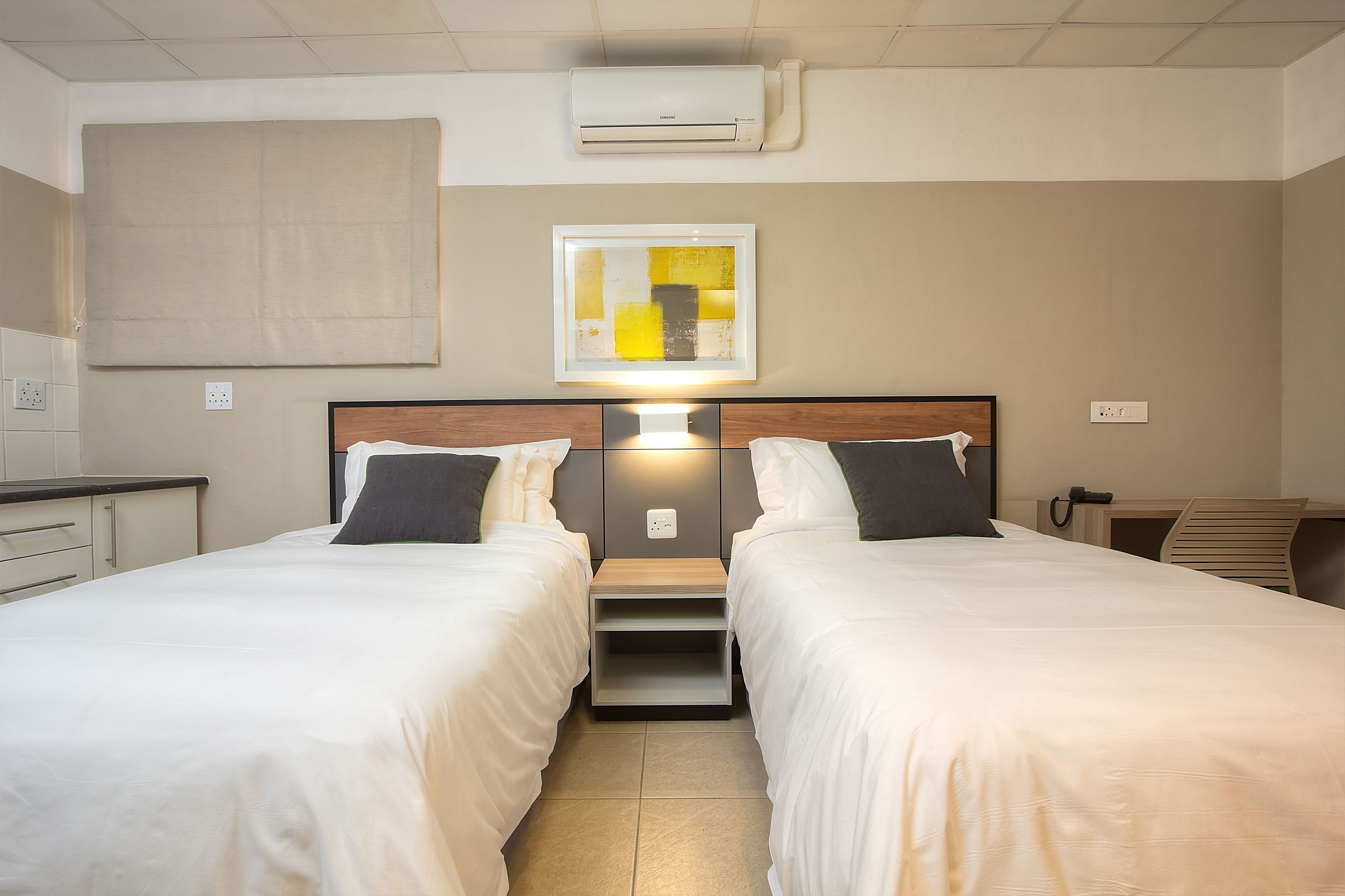 A hotel room with two beds, a bedside table, a nightstand, a wall-mounted air conditioner, a framed abstract artwork, and a desk with a chair.