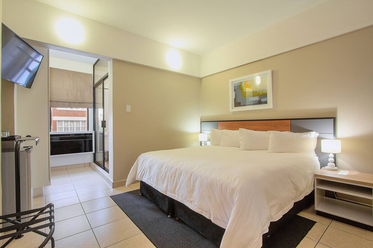 A hotel room with a bed, bedside table, lamp, TV, and a view of the outside.
