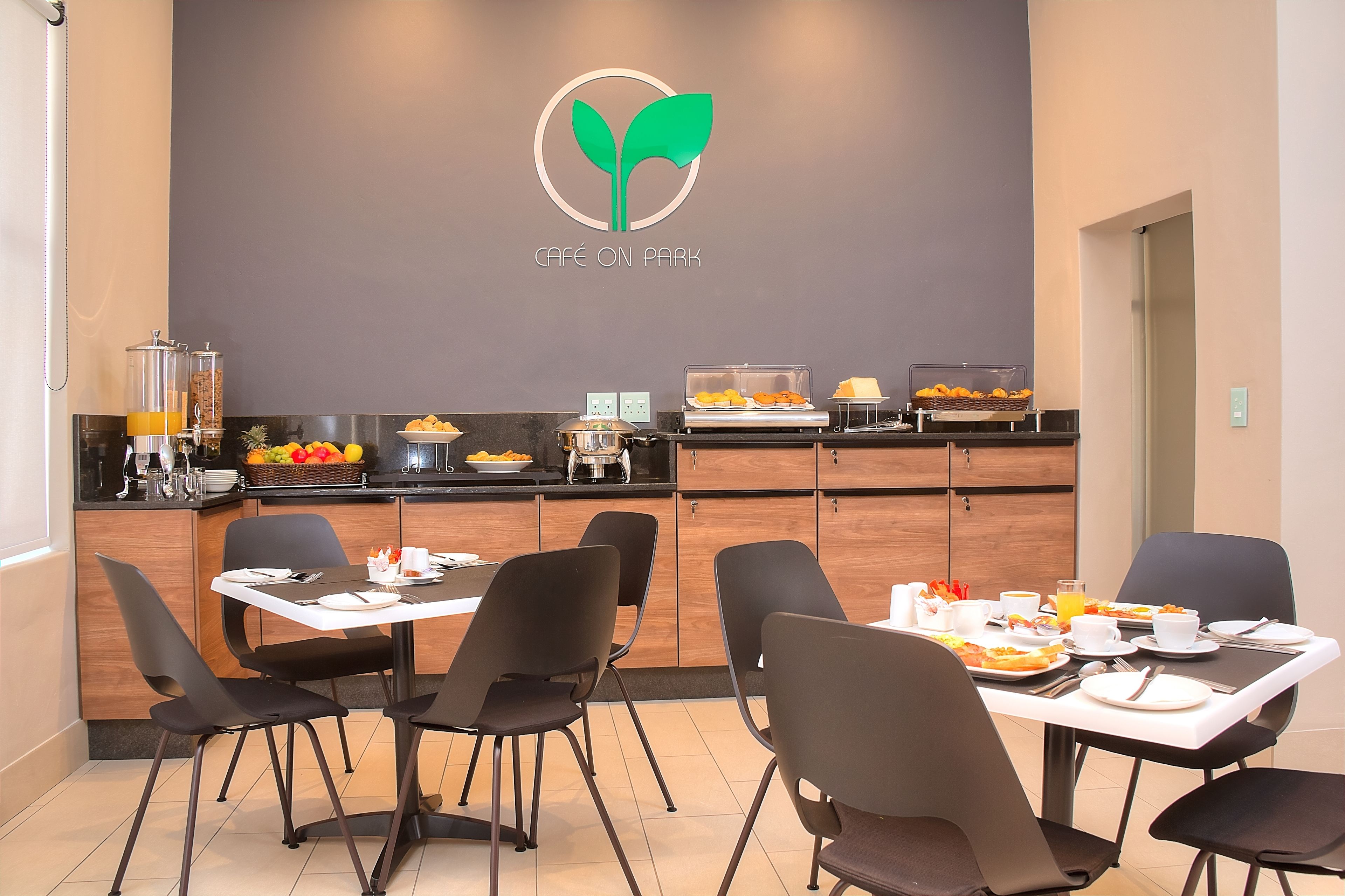 A café with a modern design, featuring a logo on the wall, a buffet with various food items, and black chairs around a table.