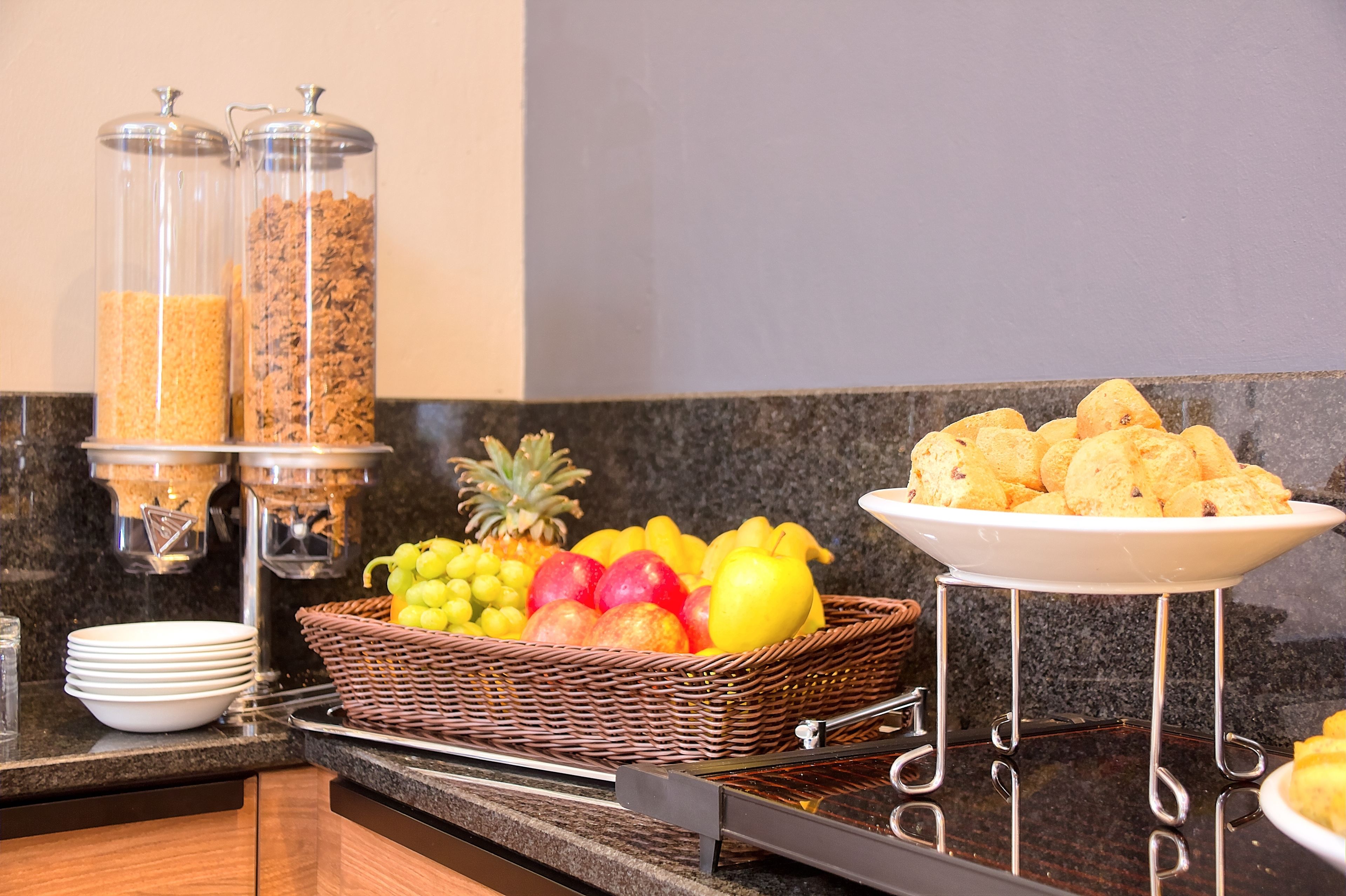A buffet setup with a fruit basket, cereal containers, and a plate of pastries.