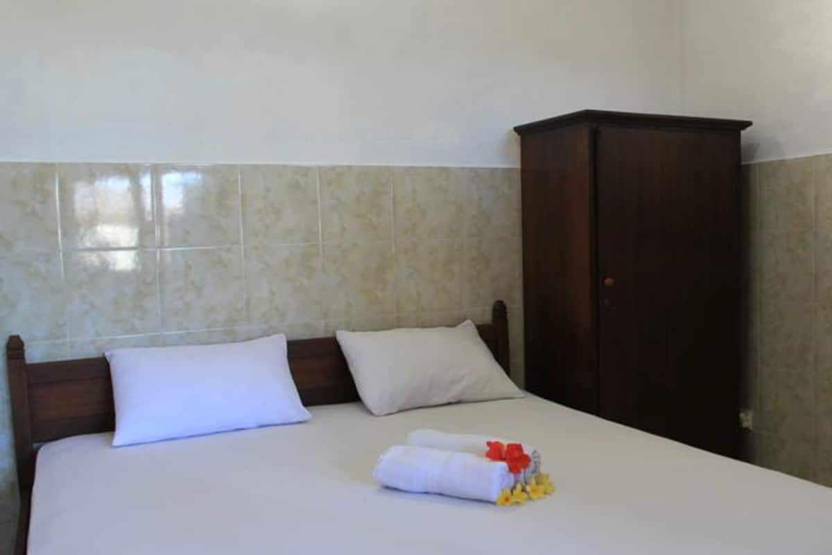 A hotel room with a bed, two pillows, a wooden wardrobe, and a neatly made bed with a towel and flowers.