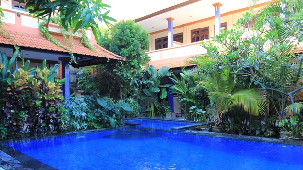 A swimming pool surrounded by lush greenery and a building with a tiled roof. - Losmen Cempaka 3 (Kuta)