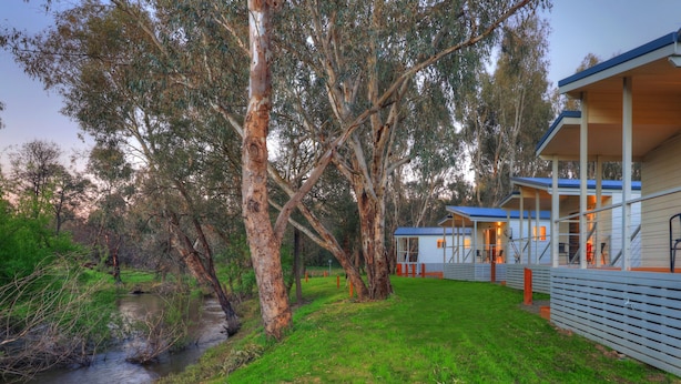 A cabin with a covered porch, a small outdoor seating area, and a view of a river. - Yea Riverside Caravan Park (Yea)