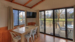A dining area with a wooden table and chairs, a flat-screen TV mounted on the wall, and large sliding glass doors leading to an outdoor patio. - Yea Riverside Caravan Park (Yea)