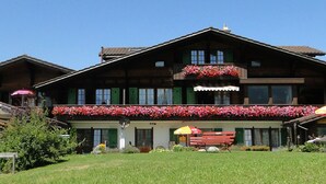 A traditional wooden chalet with a balcony adorned with flowers, a covered outdoor seating area, and a well-maintained garden. - Hotel Chalet Bergblick (Aeschi bei Spiez)