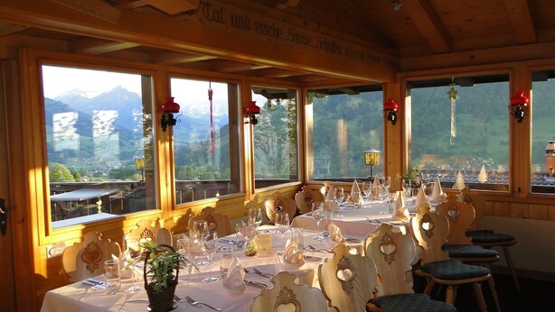 A dining area with a table set for a meal, large windows with a mountain view, and wooden interior. - Hotel Chalet Bergblick (Aeschi bei Spiez)