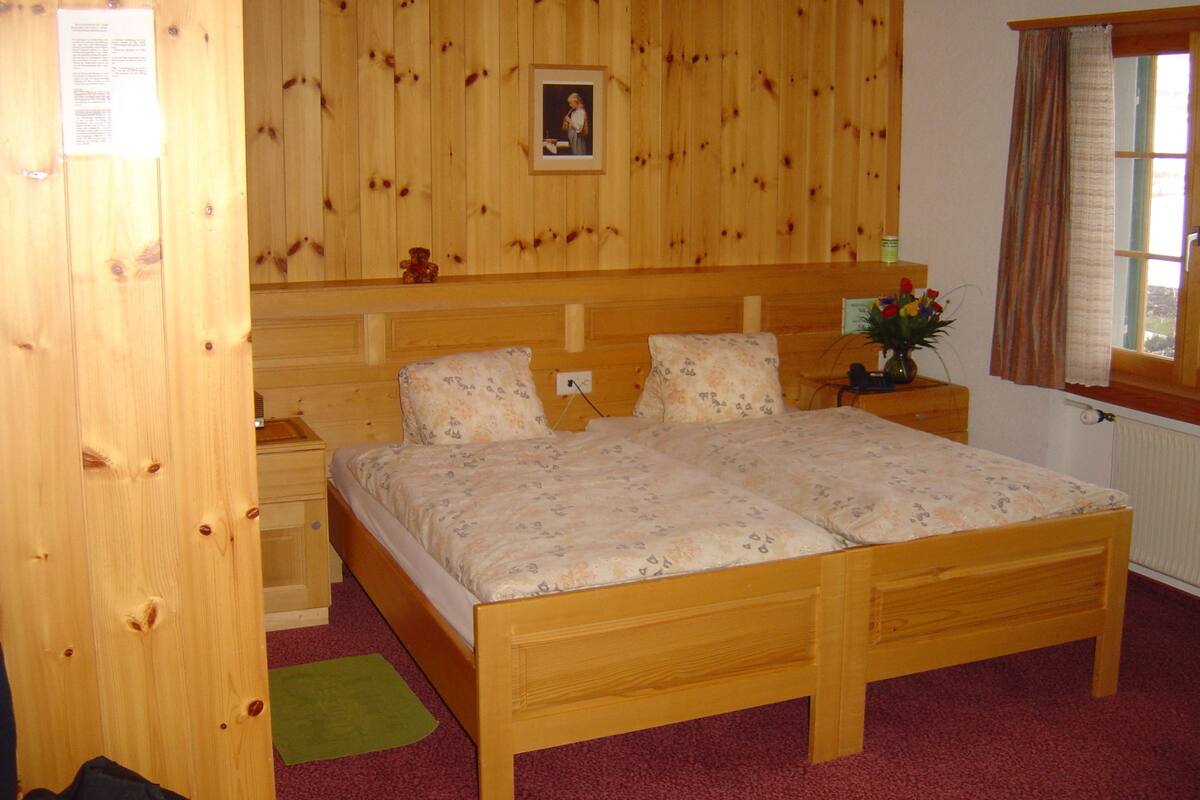 A wooden-paneled bedroom with a double bed, a nightstand, a green rug, and a window with curtains.