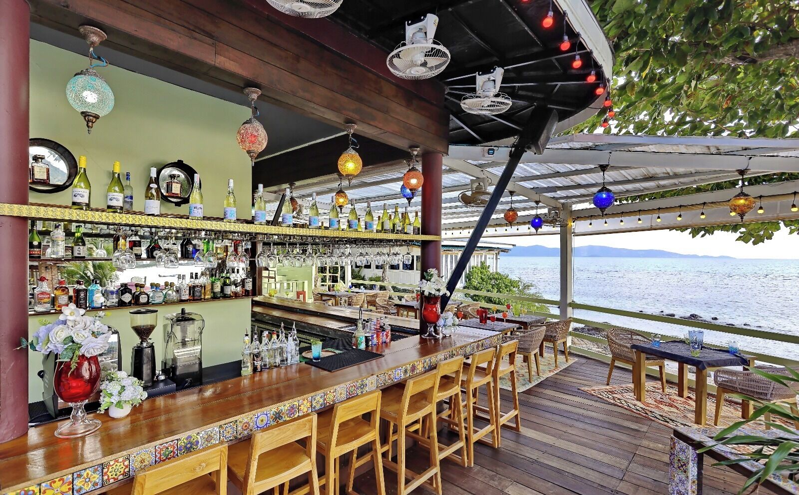 An outdoor bar area with a wooden deck, bar stools, and a view of the sea.