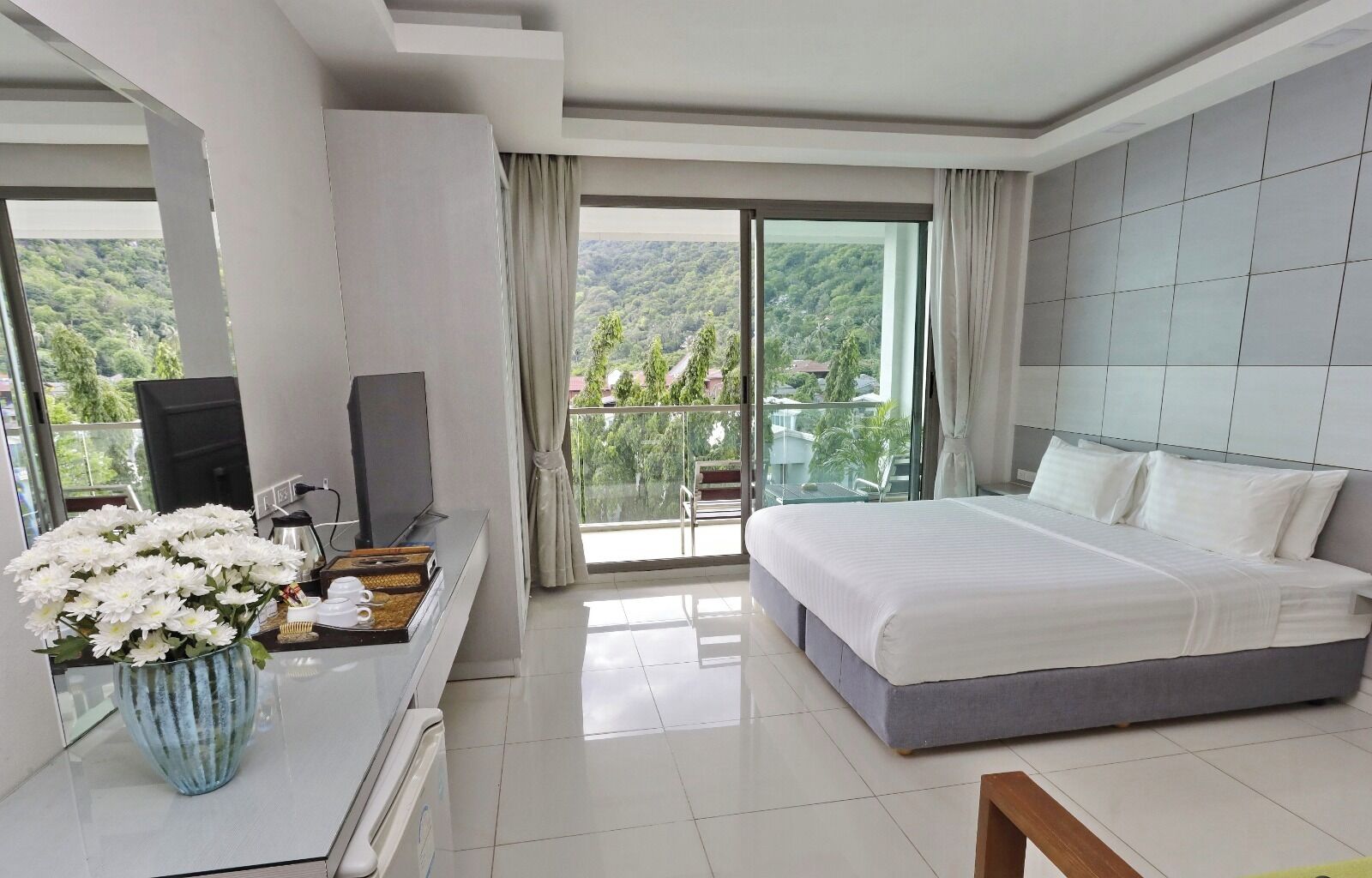 A hotel room with a bed, a TV, a desk with a vase of flowers, and a balcony with a view of the hills.