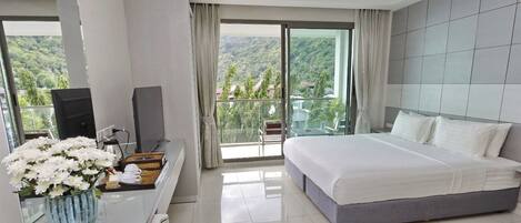 A hotel room with a bed, a TV, a desk with a vase of flowers, and a balcony with a view of the hills.