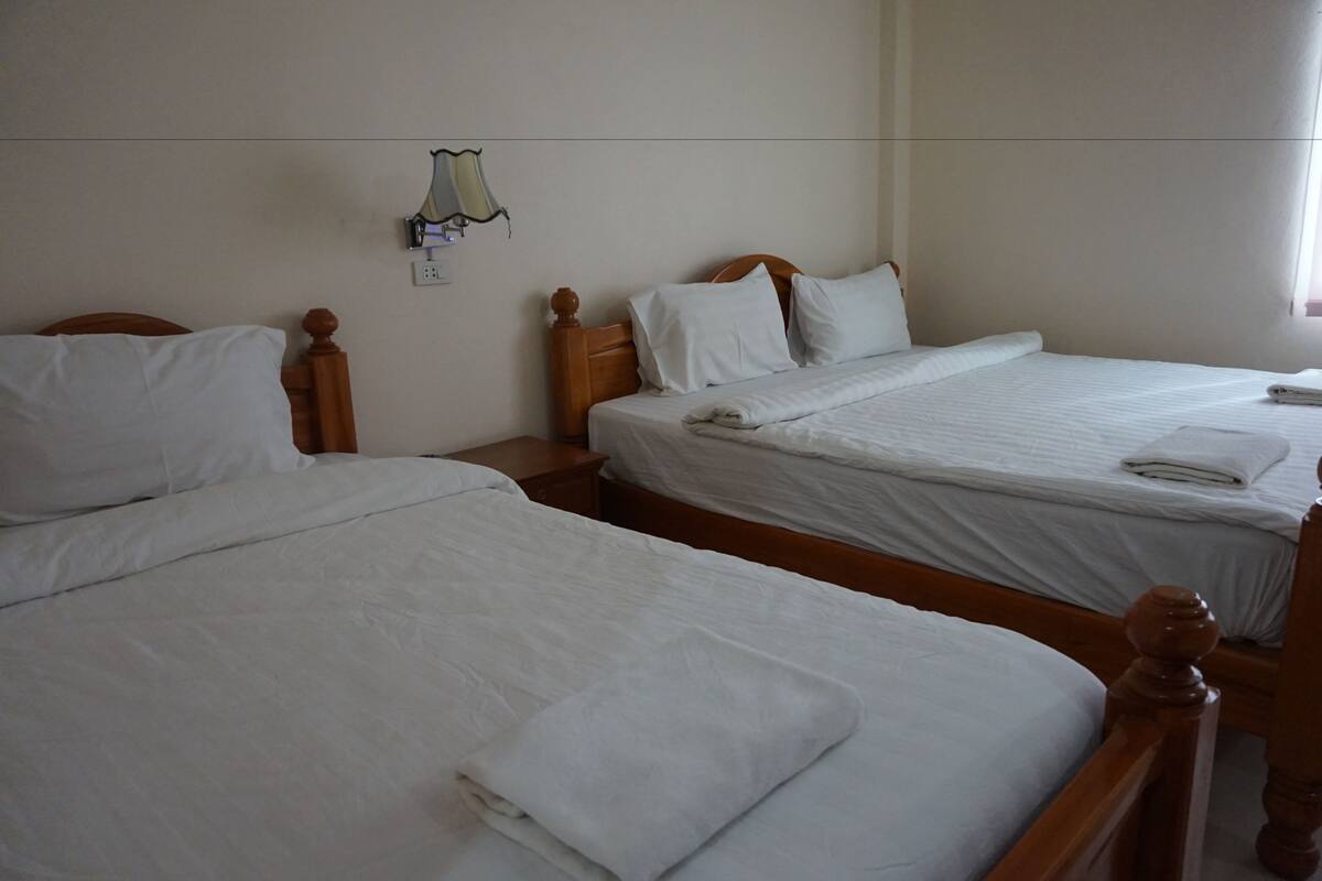Habitación de hotel con dos camas individuales, cabeceros de madera, ropa de cama blanca, una mesita de noche pequeña y una lámpara fijada a la pared.