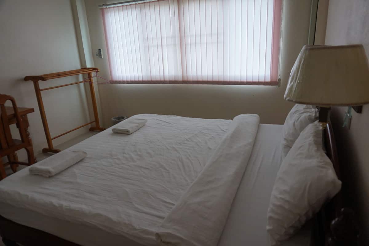 Una cama bien hecha con sábanas blancas, una silla de madera, una mesita de noche con una lámpara y una ventana con persianas.