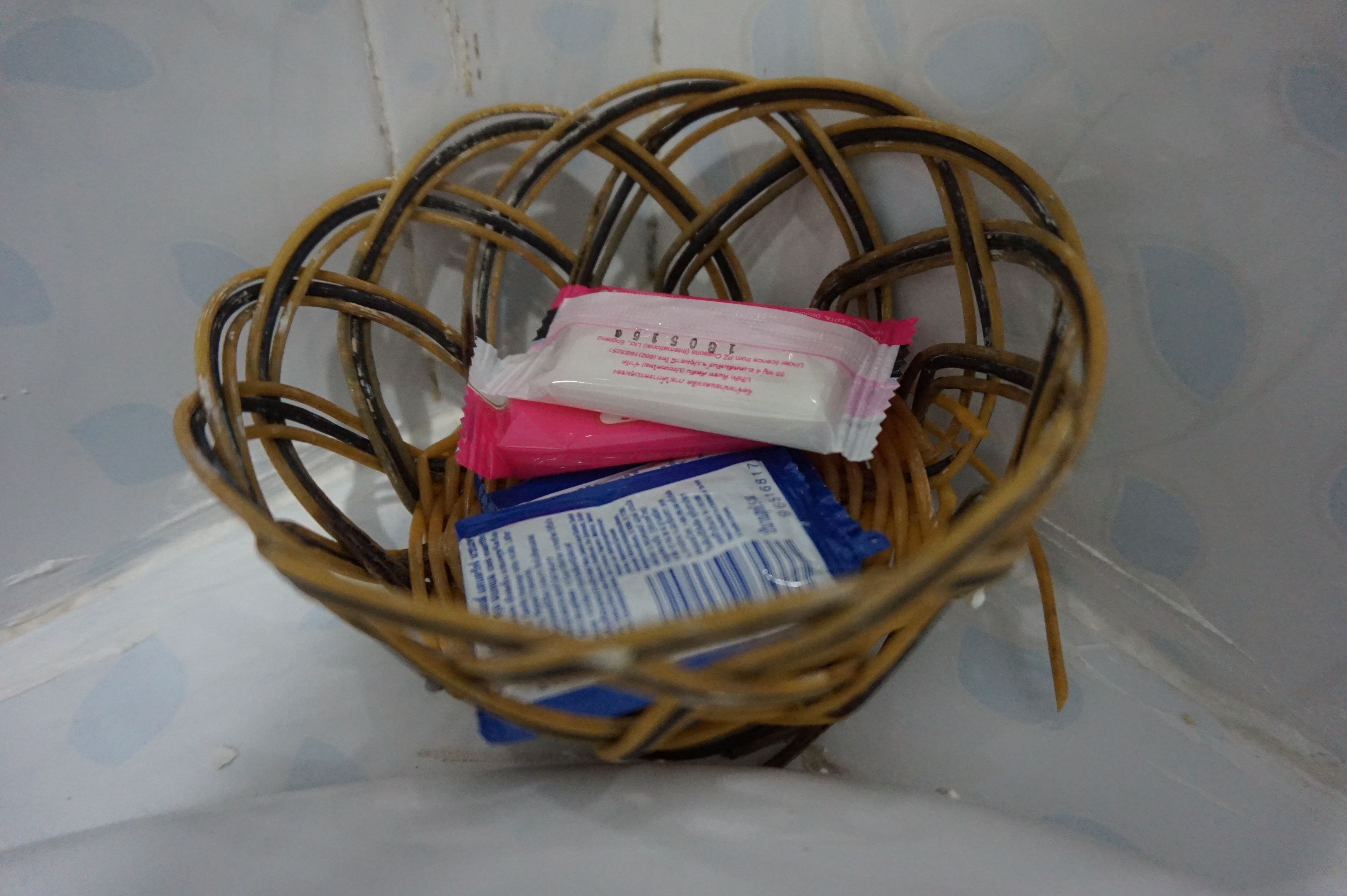 A wicker basket containing a pink and a blue package, possibly toiletries, in a room with a patterned wall.