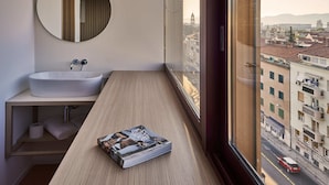 Un bagno moderno con uno specchio rotondo, un lavandino bianco e un mobile contenitore in legno. Una rivista è posata sul mobile, e la stanza offre una vista sulla città attraverso una grande finestra.