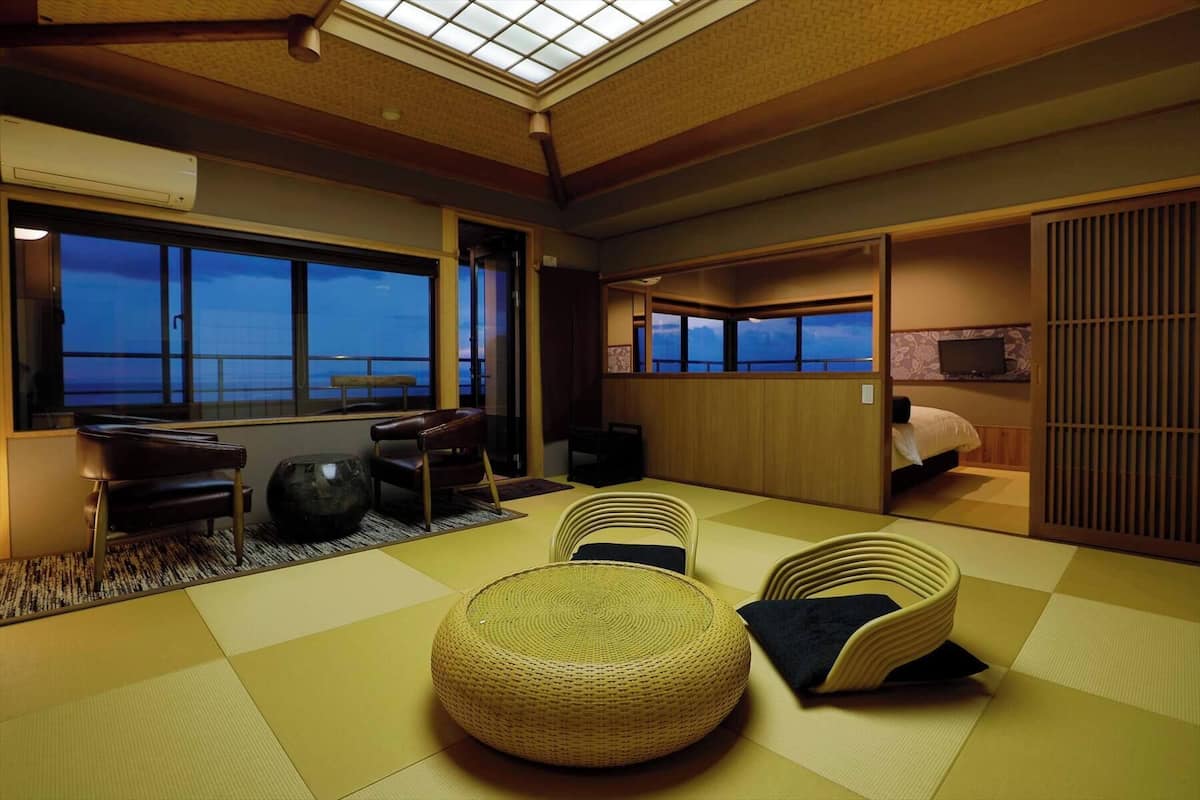 A traditional Japanese room with tatami flooring, a low table, a cushioned chair, and a bed with a view of the sea.