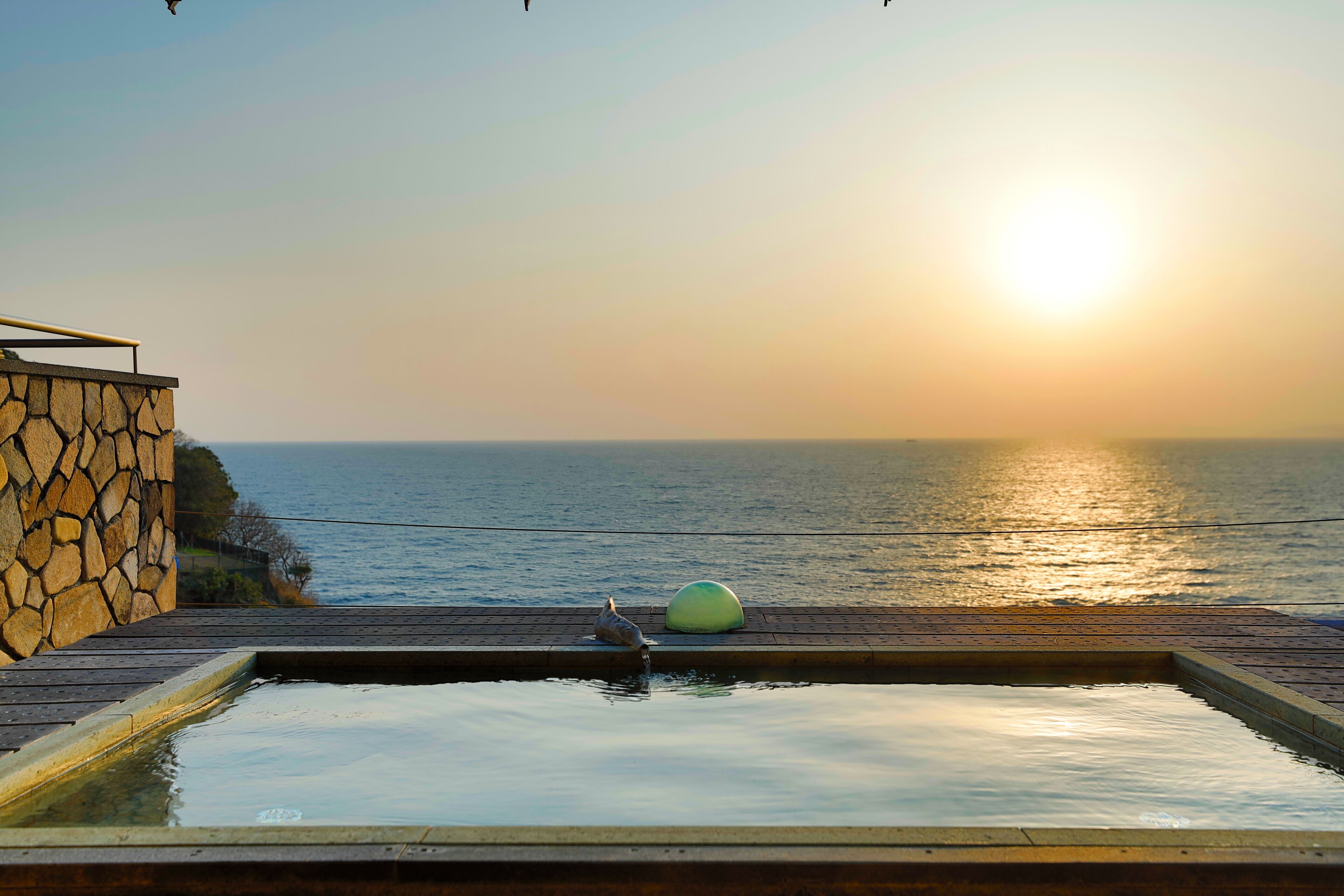 A stone wall, a wooden deck, and a swimming pool with a sunset view.