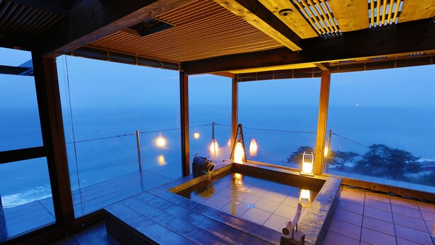 A modern interior with large windows overlooking a body of water, a wooden ceiling, and a small seating area with a table and chairs. - Hotel Isaba (Numazu)