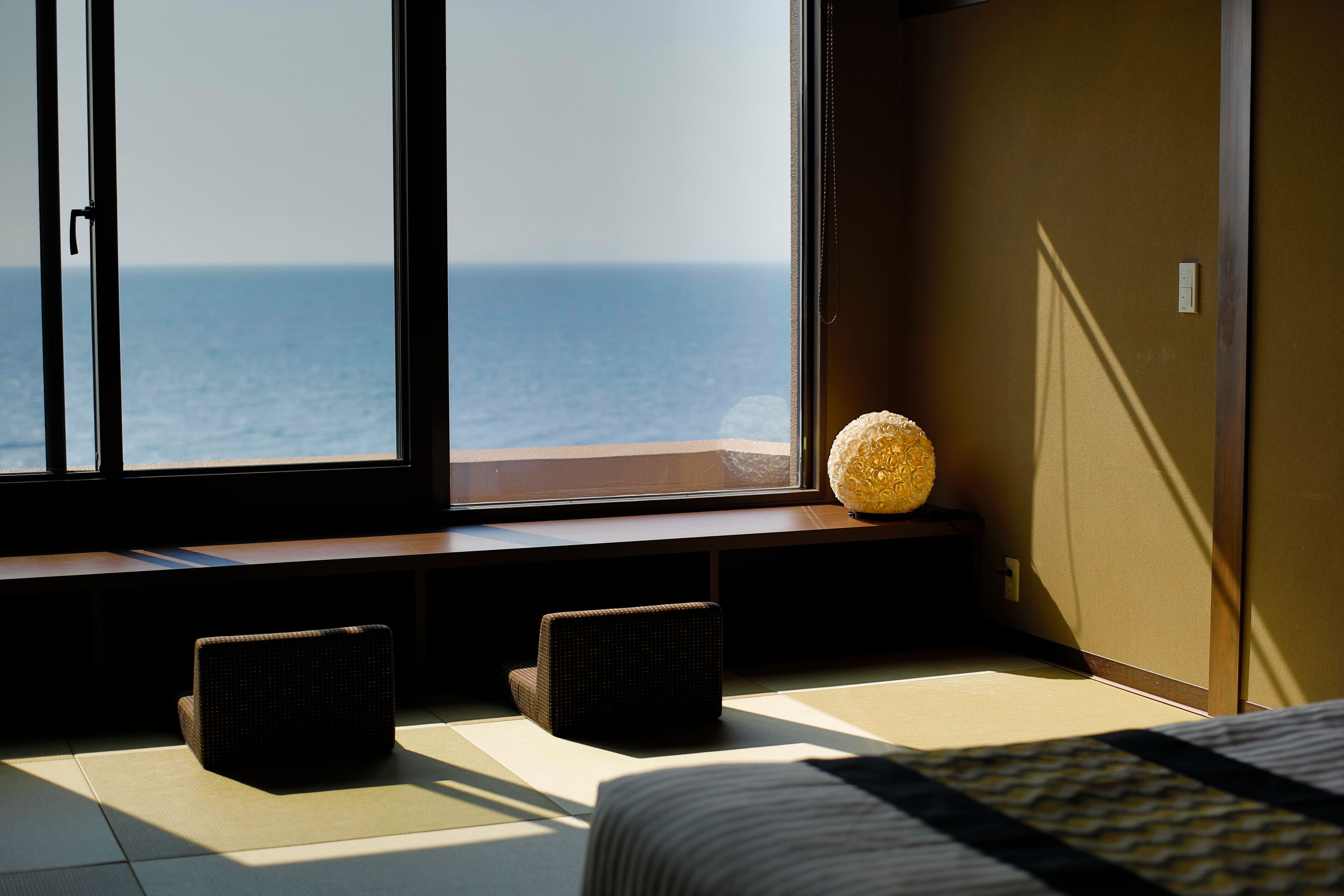 A hotel room with a large window overlooking the sea, a bed with a patterned bedspread, and two dark-colored chairs.