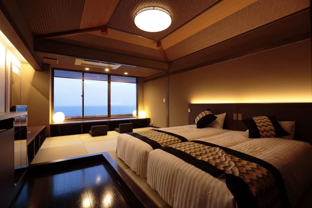 A hotel room with two beds, a large window with a view, a ceiling light, and a bedside table.