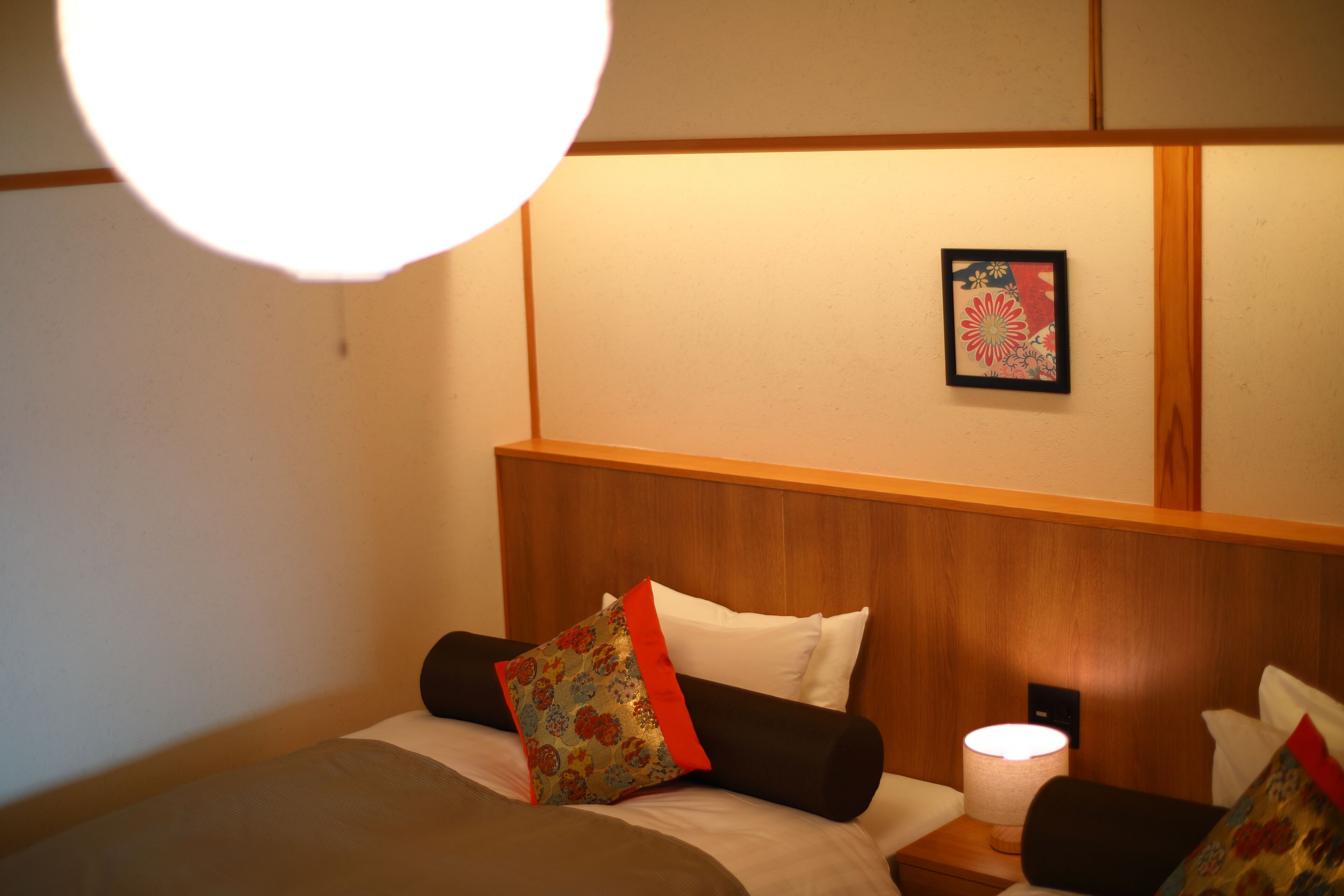 Quarto de hotel com cabeceira de madeira, cama com roupa de cama branca e castanha, mesa de cabeceira com abajur e quadro pendurado na parede.