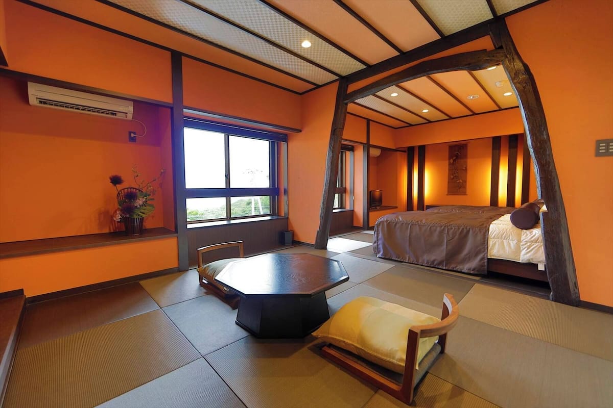 A traditional Japanese-style room with tatami flooring, a low table, a bed, and a large window.
