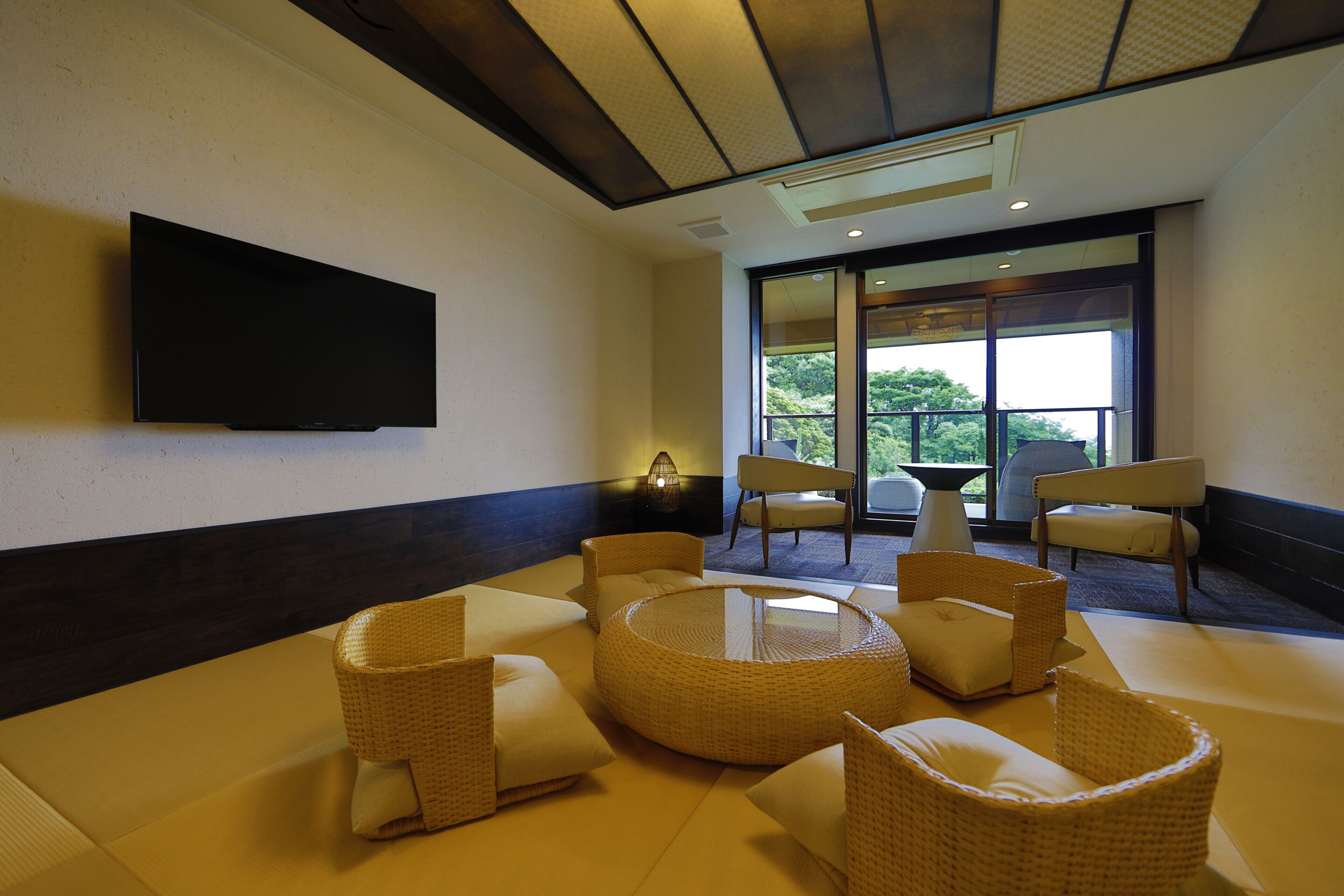 A modern lounge area with wicker furniture, a flat-screen TV, and large windows offering a view of greenery.