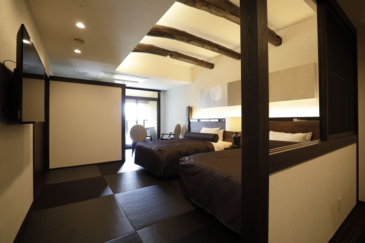 A hotel room with two beds, a sitting area with a chair, a flat-screen TV mounted on the wall, and a large window providing natural light.