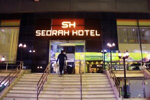 El exterior del Hotel SH Serrah, con un llamativo letrero iluminado, una escalera que conduce a la entrada y el reflejo del hotel en el fachada de vidrio.