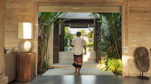 A man walking through a wooden entrance with a glass door, surrounded by tropical plants and a stone sculpture.