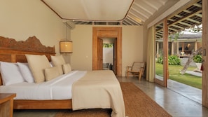 Un dormitorio con cabecera de madera, cama con ropa blanca, mesita de noche, lámpara y vistas a un jardín a través de una puerta de cristal.