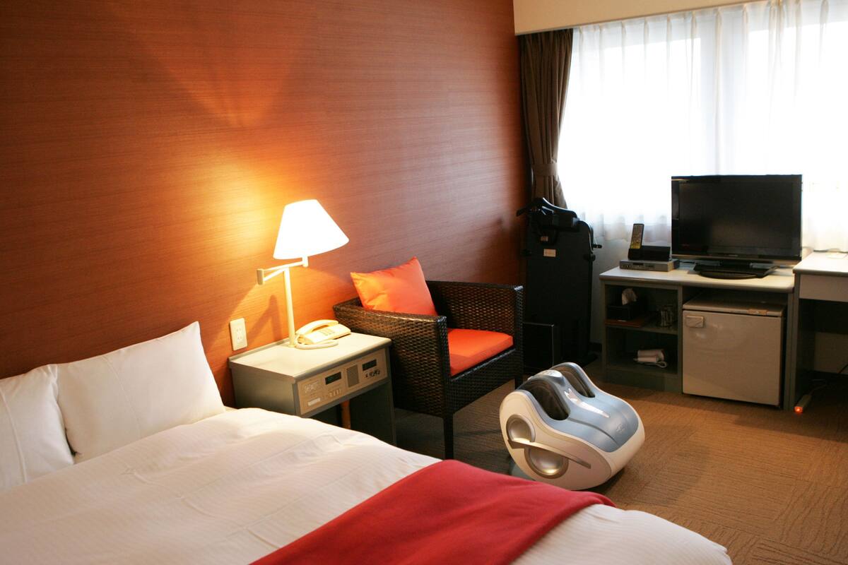 A hotel room with a bed, a chair, a desk with a television, a small refrigerator, and a massage chair.