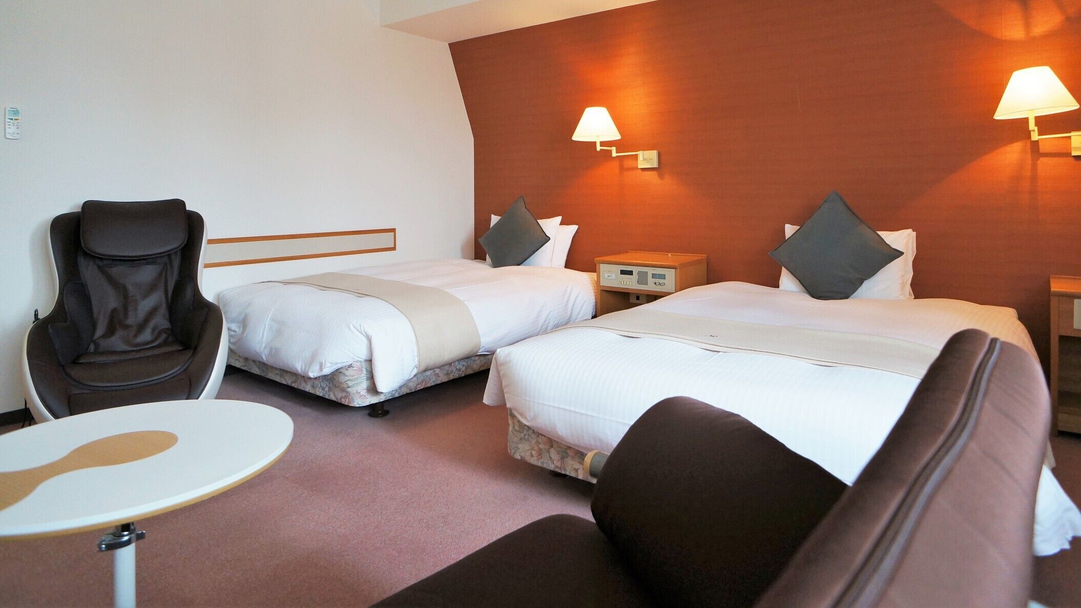 Habitación de hotel con dos camas, una silla, una mesita pequeña y una mesita de noche con lámpara.