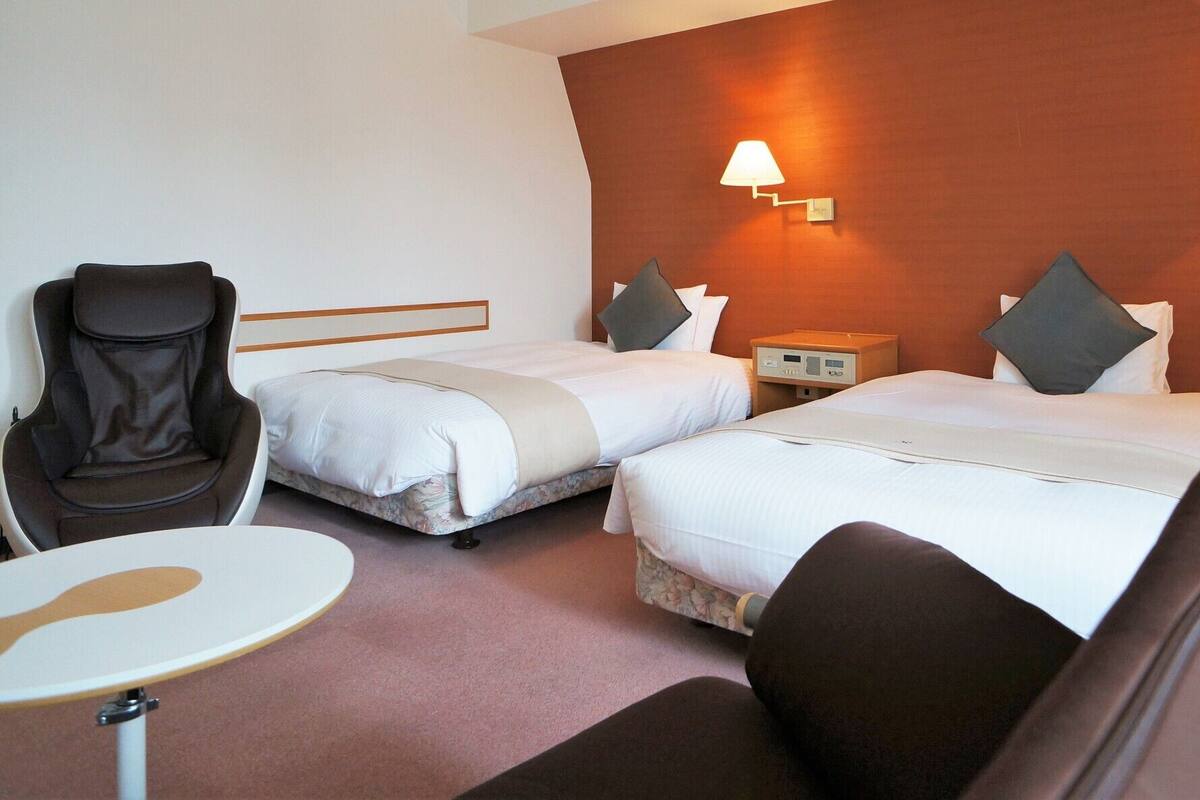 A hotel room with two beds, a chair, a small table, and a nightstand with a lamp.