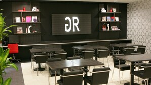A modern conference room with multiple tables and chairs, a large screen displaying 'OR', and shelves with books and decorative items.