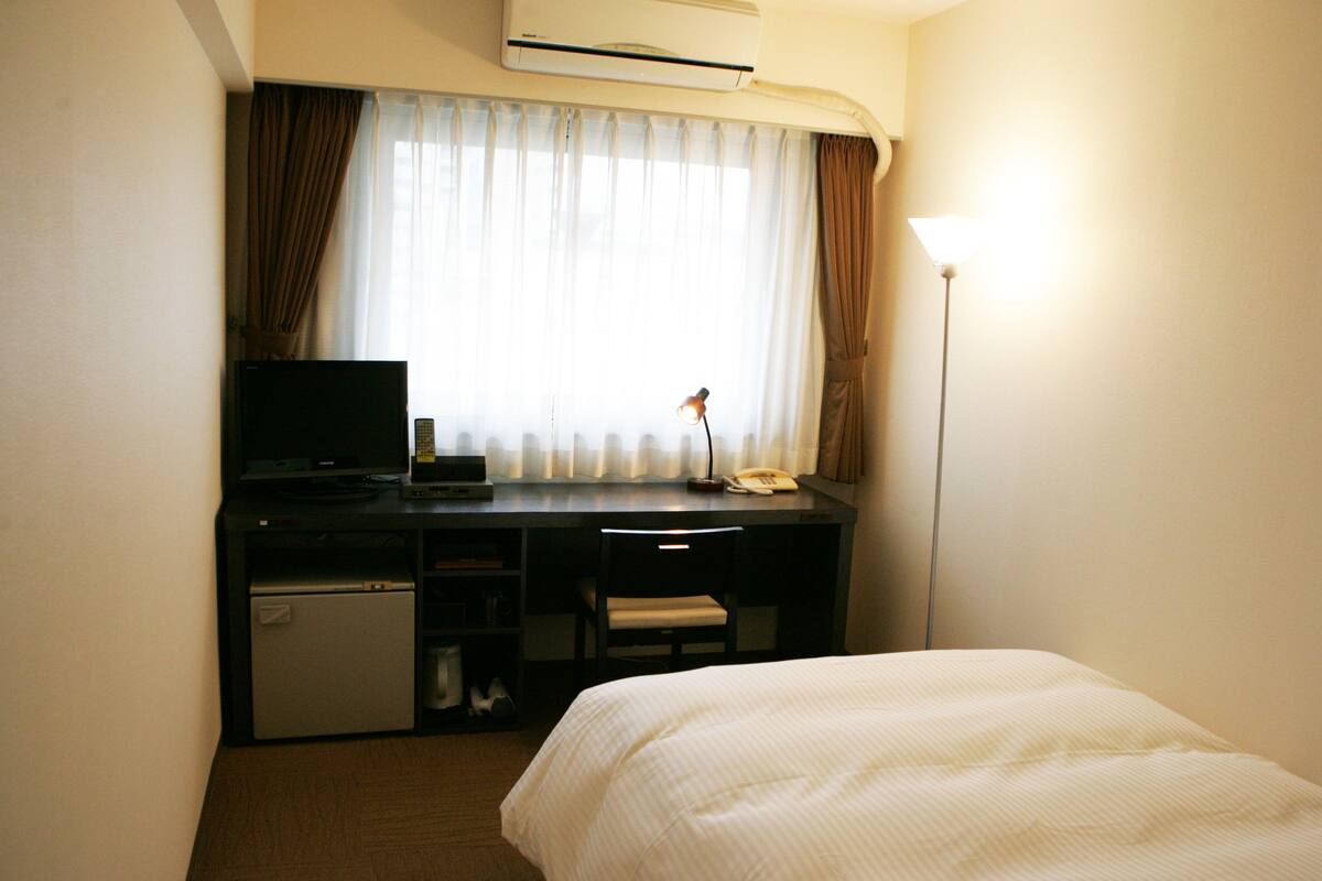 A hotel room with a bed, a desk with a television, a small refrigerator, and a lamp.