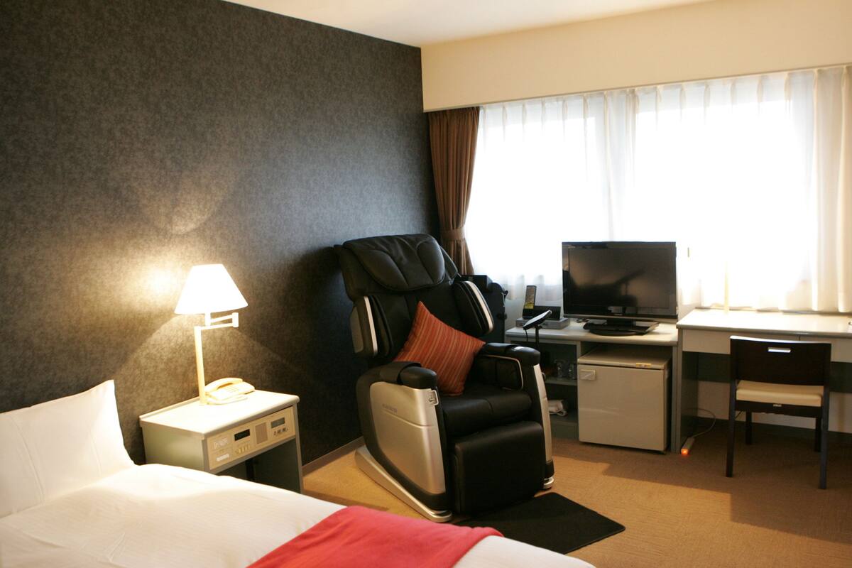 A hotel room with a bed, massage chair, desk, TV, and a small refrigerator.