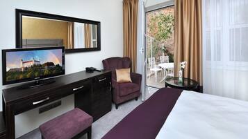 Habitación de hotel con cama, televisor, escritorio con cajones, silla, mesita y balcón con vista a un edificio y vegetación.