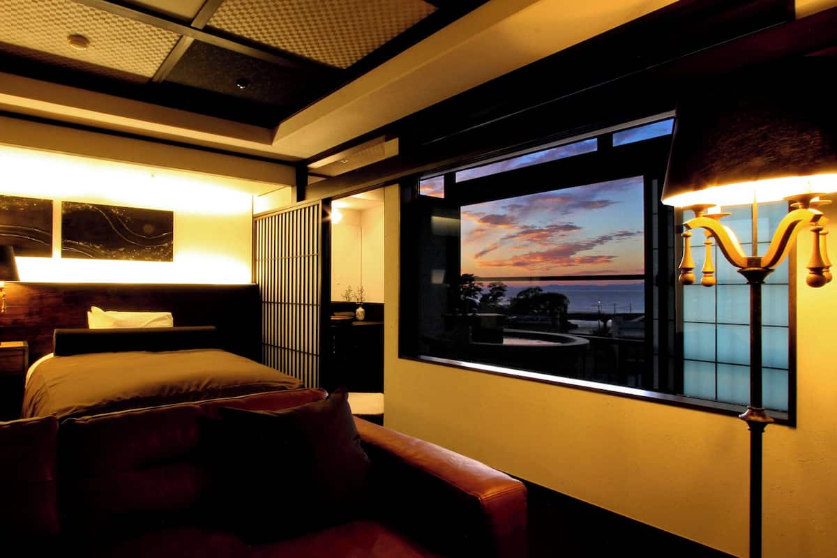 A hotel room with a bed, bedside tables, a lamp, a window with a sunset view, and a decorative wall panel.