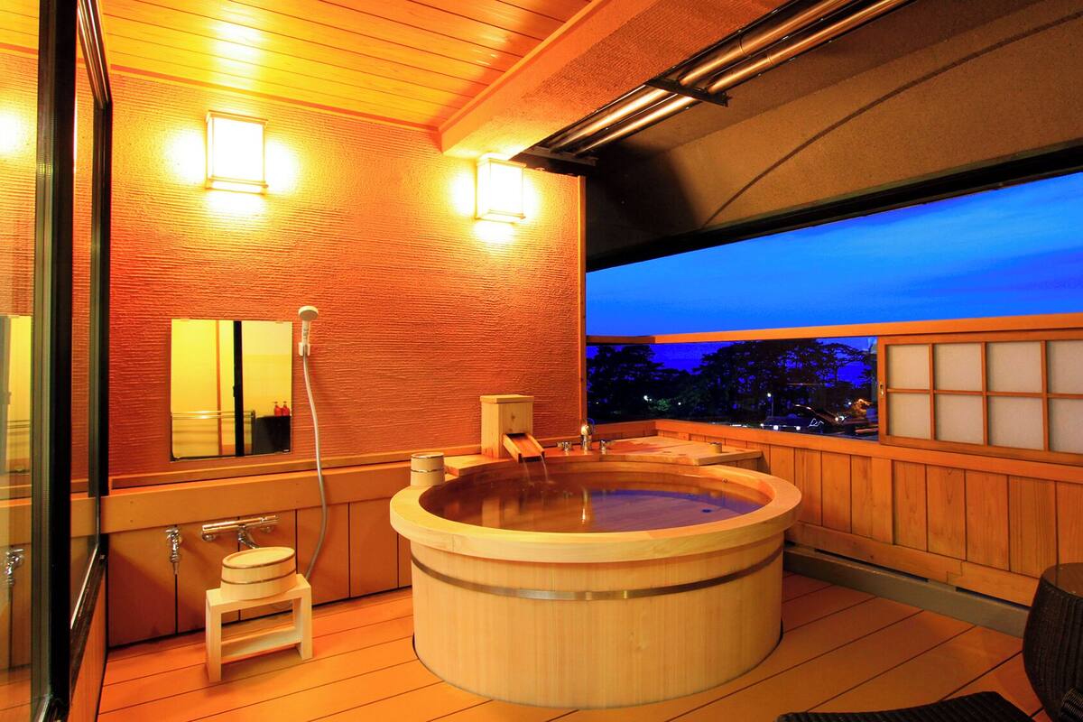 A wooden spa with a hot tub, a small wooden stool, and a view of the city at dusk.