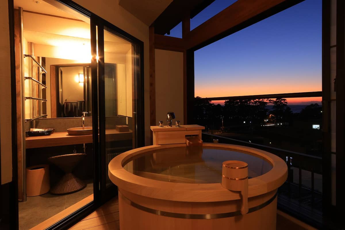 A modern bathroom with a glass-enclosed shower, a wooden bathtub, and a view of the sunset.