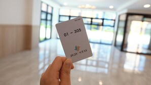 A hand holding a card with "01 - 305" and "JEJU STAY" text, in a bright indoor space with large windows. - Jeju Stay in Seongsan Hotel (Seogwipo)