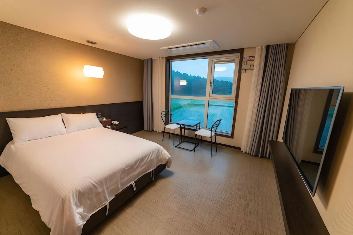 A hotel room with a bed, a desk, a chair, a large window with a view of the sea, and a mirror.