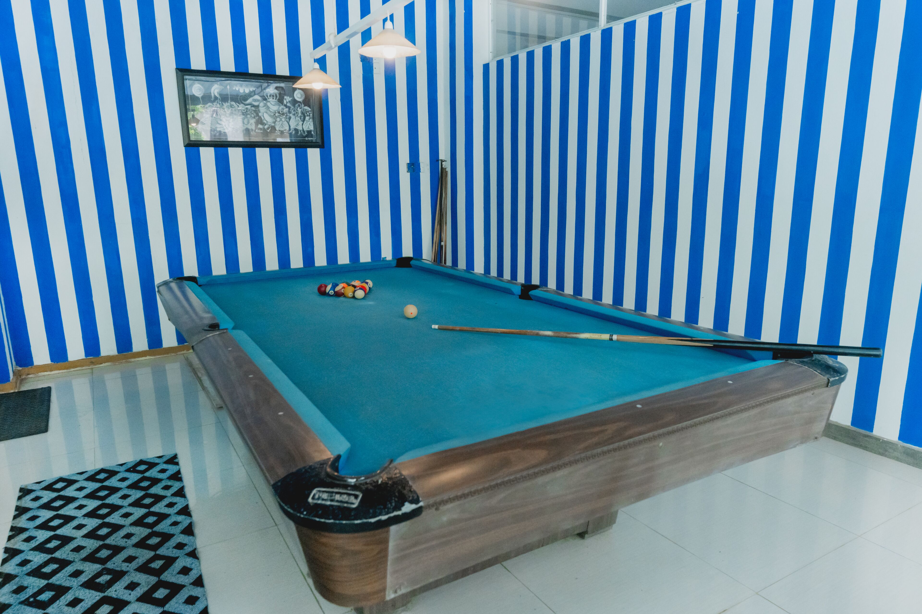 Una mesa de billar con superficie de fieltro azul, rodeada de paredes a rayas azules y blancas, y un cuadro en la pared.