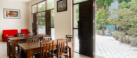 A dining area with wooden tables and chairs, a red sofa, and large windows offering a view of the outdoors.