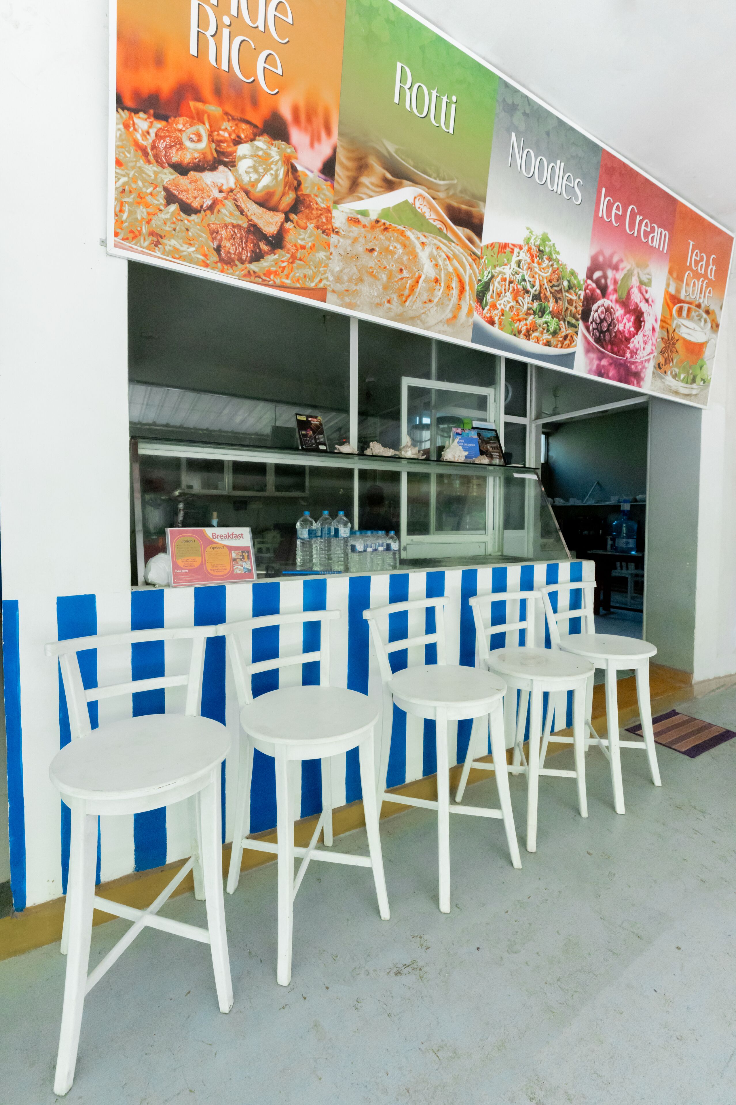 Un petit stand de restauration proposant un menu affiché sur un panneau, avec des plats comme du riz, des galettes, des nouilles, de la glace, ainsi que du thé et du café. Quatre tabourets blancs sont placés devant le stand.