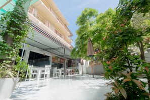 An outdoor patio area with white furniture, a covered seating area, and a building with a balcony in the background.