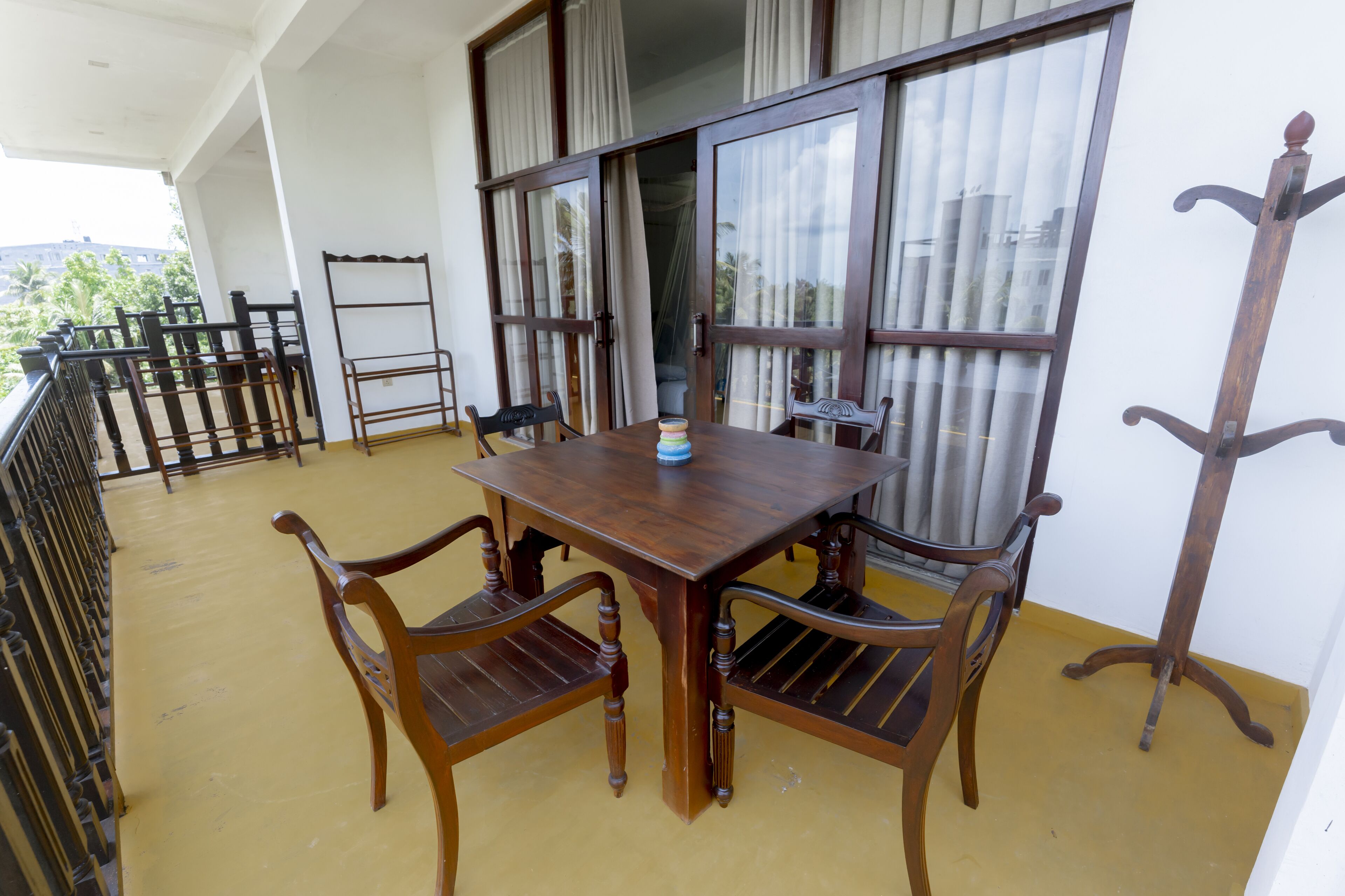 Una mesa de madera con cuatro sillas en un balcón con piso amarillo, ventanas amplias con cortinas y barandilla de metal.