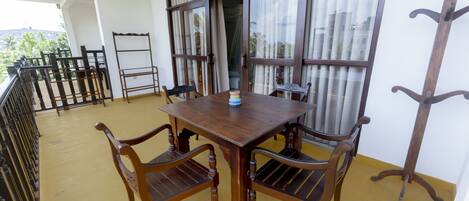 Una mesa de madera con cuatro sillas en un balcón con piso amarillo, ventanas amplias con cortinas y barandilla de metal.