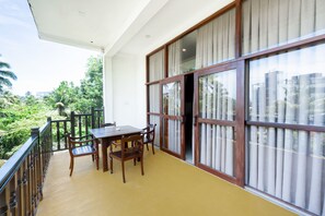 A balcony with a wooden table and chairs, large windows with curtains, and a view of greenery and buildings.